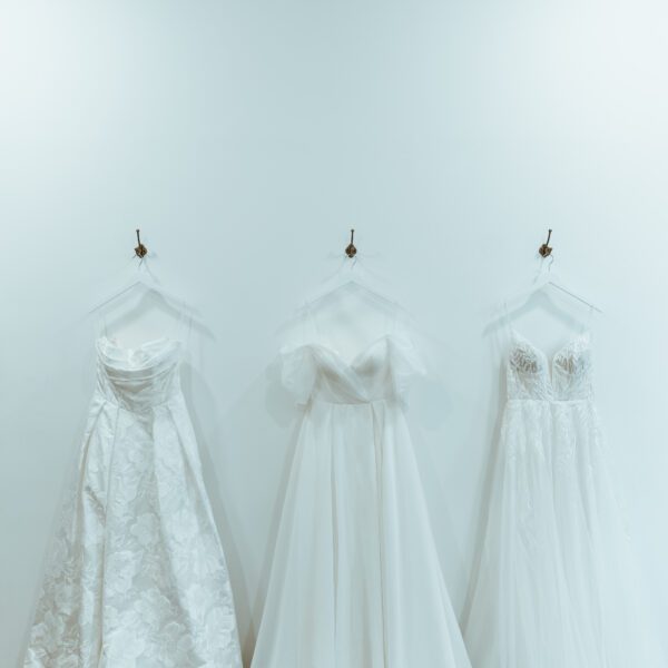 A wide lens shot of 3 designer wedding dresses hanging against a white wall on metal hooks at you are precious bridal.