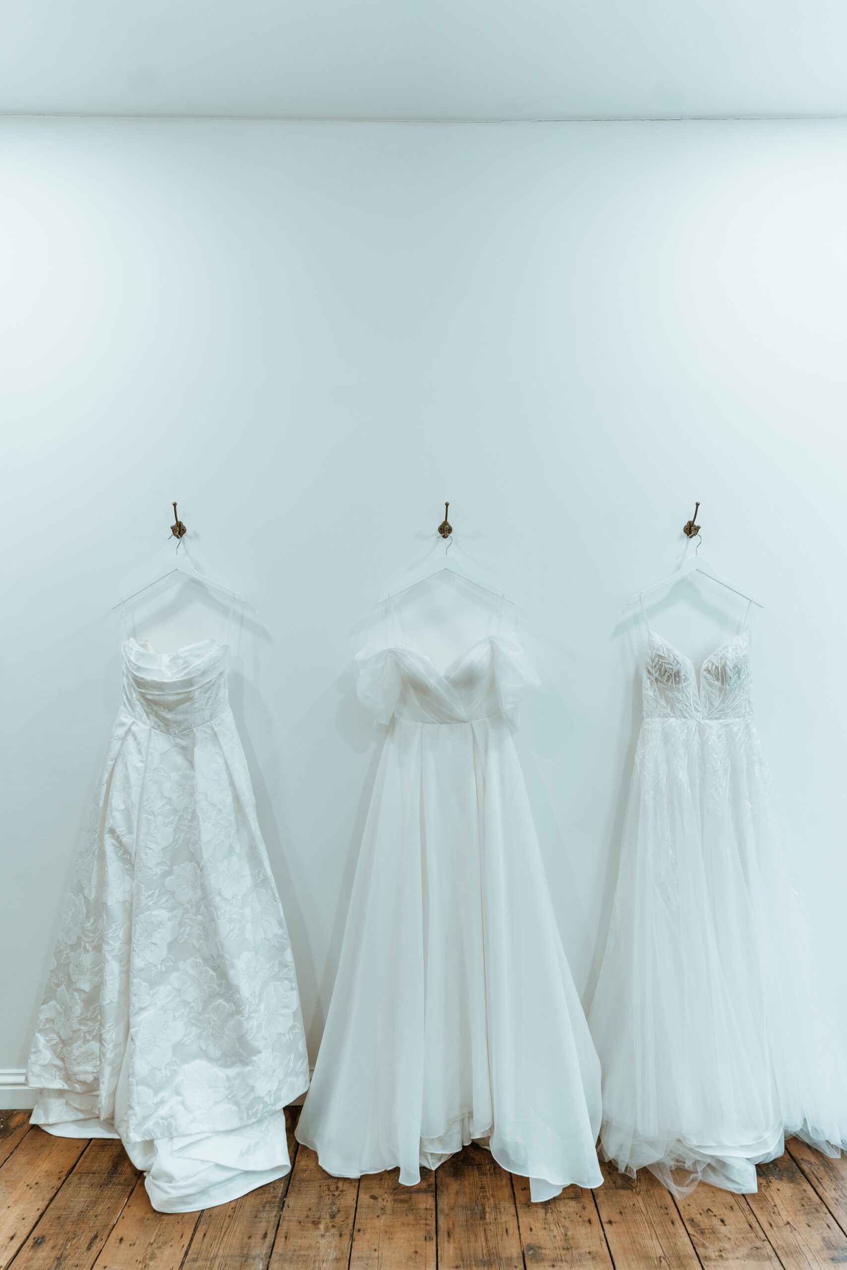 A wide lens shot of 3 designer wedding dresses hanging against a white wall on metal hooks at you are precious bridal.